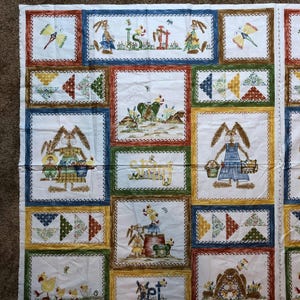 May include: A white fabric panel featuring various rectangular and square blocks with colorful illustrations. The blocks depict bunnies, butterflies, and the word "Spring". The borders are in shades of blue, green, yellow, and red.