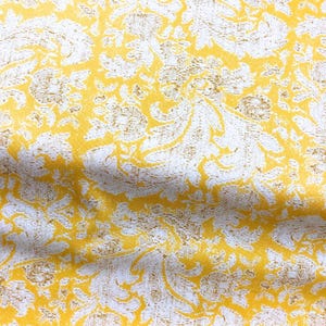 May include: Close-up of a fabric with a repeating floral pattern. The design features yellow and gold-colored floral shapes against a white background. The fabric has a textured appearance, suggesting a woven material.