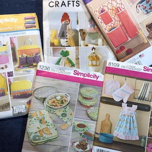 May include: A collection of Simplicity sewing pattern booklets featuring various craft projects. The patterns include designs for sewing accessories, kitchen items, and other fabric crafts. The booklets have colorful illustrations and text.