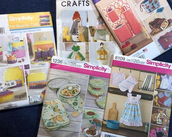 May include: A collection of Simplicity sewing pattern booklets featuring various craft projects. The patterns include designs for sewing accessories, kitchen items, and other fabric crafts. The booklets have colorful illustrations and text.
