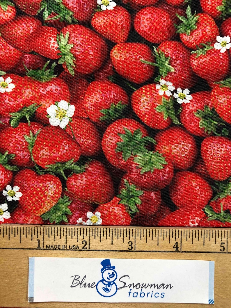 May include: Close-up of a fabric with a red strawberry print. The fabric is measured with a ruler, showing a length of 5 inches. The fabric has a label that says "Blue Snowman Fabrics".