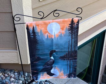 May include: A garden flag with a black and white loon on a lake with a full moon in the background. The flag is orange and blue with a forest scene.