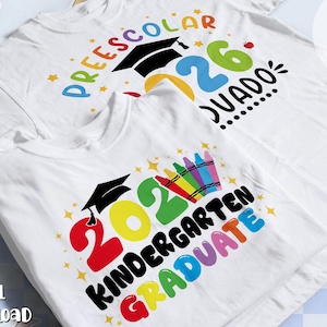 Puede incluir: Dos camisetas blancas con diseños gráficos coloridos. Una muestra el texto PREESCOLAR GRADUADO con un birrete negro y el año 2026. La otra presenta el texto KINDERGARTEN GRADUATE con un birrete negro, el año 2026 y una fila de lápices de colores.