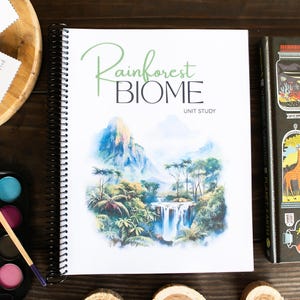 May include: A spiral-bound "Rainforest Biome Unit Study" book with a watercolor illustration of a waterfall and mountains. A watercolor paint set and a book titled "The Wonderous Workings of Planet Earth" are also visible.