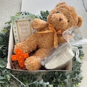May include: A gift box filled with baby items, including a plush brown teddy bear with a yellow ribbon, a book titled "Baby's First Book of Prayers", a white and gray teething toy, and an orange pop-it toy shaped like a cross.