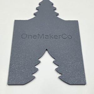 May include: A gray, tree-shaped item with the text "OneMakerCo" embossed on the surface. The item has a textured appearance and a cutout at the bottom, also in the shape of a tree. The top of the item is also shaped like a tree.