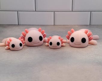 May include: Four pink axolotl figurines with black eyes and pink gills are arranged on a gray surface. The axolotls vary in size, with the largest in the center and the smallest on the left.