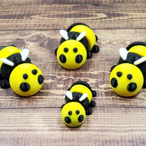 May include: Five yellow and black bee figurines with white antennae are arranged on a wooden surface. The bees have black eyes and are in a resting position. The background is a brown wooden surface.