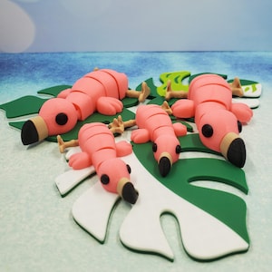 May include: Five pink flamingo figurines with black eyes and beaks, lying on a green and white leaf-shaped surface.