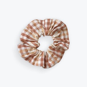 May include: A brown and cream gingham scrunchie. The fabric is gathered around a central opening, creating a ruffled effect. The pattern is a classic check design, with alternating squares of brown and cream.
