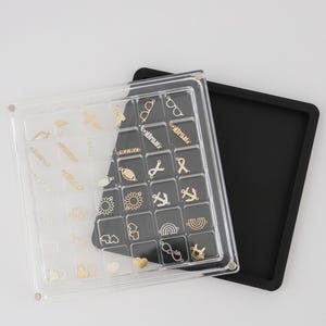 May include: A clear plastic organizer with a black base, filled with small gold-colored charms. The charms include shapes like eyeglasses, scissors, anchors, and hearts. The organizer is open, revealing the charms.