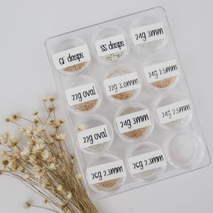 May include: A clear plastic storage container with 10 small containers filled with gold and silver jump rings. Each container has a label with the size and gauge of the jump rings. The labels read: GF dasps, SS dasps, 24g 3mm, 22g oval, 22g 3.0mm, 24g 2.5mm, 22g oval, 24g 3mm, 24g 2.5mm, 2cg 2.3mm, 2cg 2.3mm.