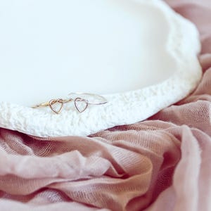 May include: Two delicate rings with heart-shaped details. One ring is gold and the other is silver. The rings are displayed on a white surface with a pink fabric background.