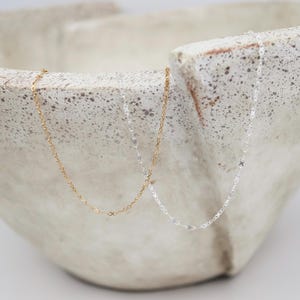 May include: Two delicate necklaces, one gold-toned and one silver-toned, are displayed against a neutral, textured background. The necklaces feature a fine chain with small, decorative elements. The gold necklace is on the left, and the silver necklace is on the right.