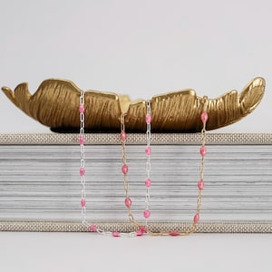 May include: Three necklaces with pink gemstone beads. One necklace is silver, and the other two are gold. The necklaces are displayed in front of a gold leaf-shaped decorative bowl, resting on a book.