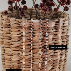 May include: Four gold chains with different colored beads hanging from a wicker basket. The chains are labeled "Harvest Plaid" and "Sweater Weather".
