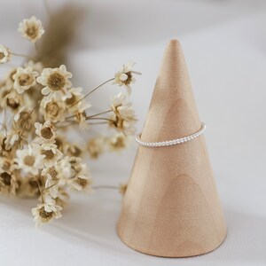 May include: A delicate silver ring with a beaded design sits on a wooden ring display stand. The stand is a light brown color and is shaped like a cone. Dried white flowers are in the background.