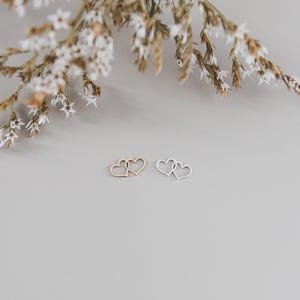 May include: Two heart-shaped pendants, one gold-toned and one silver-toned, interlocked. Delicate white flowers and foliage are in the background, set against a neutral gray backdrop. The pendants are small and likely intended for jewelry.