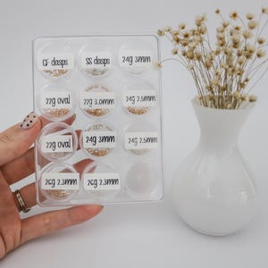 May include: A clear plastic container with 10 small compartments, each containing different sizes and styles of gold jump rings. The compartments are labeled with the weight and size of the jump rings.  CF dasps, SS dasps, 24g 3mm, 22g oval, 22g 3.0mm, 24g 2.5mm, 22g oval, 24g 3mm, 24g 2.5mm, 2cg 2.3mm, 2cg 2.3mm.