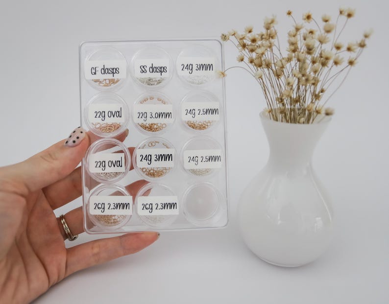 May include: A clear plastic container with 10 small compartments, each containing different sizes and styles of gold jump rings. The compartments are labeled with the weight and size of the jump rings.  CF dasps, SS dasps, 24g 3mm, 22g oval, 22g 3.0mm, 24g 2.5mm, 22g oval, 24g 3mm, 24g 2.5mm, 2cg 2.3mm, 2cg 2.3mm.