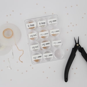 May include: A clear plastic container with small compartments filled with different sizes and types of gold beads. The container is labeled with the bead sizes and types, such as "CF dasps", "SS dasps", "22g oval", "24g 3mm", and "24g 2.5mm". A pair of black wire cutters is also in the image.