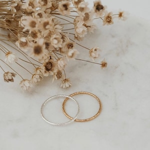 May include: Two delicate rings, one silver and one gold, with a textured design, are displayed on a marble surface. Dried flowers in shades of beige and brown are arranged in the background, adding a touch of natural elegance.