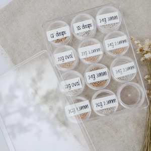 May include: A clear plastic storage box with 10 small containers filled with different types of gold and silver jump rings. The containers are labeled with the size and gauge of the jump rings.  The labels include: GF dasps, SS dasps, 22g oval, 22g 3.0mm, 24g 3mm, 24g 2.5mm, 24g 3mm, 24g 2.5mm, 2cg 2.3mm, 2cg 2.3mm.