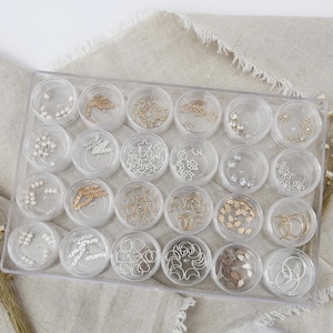 May include: A clear plastic organizer filled with small, round containers. Each container holds various decorative elements, including pearls, stars, and floral shapes in silver and gold tones. The organizer sits on a neutral-colored fabric.