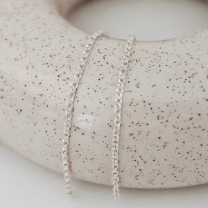 May include: A silver chain necklace with a delicate, twisted design. The chain is lying on a speckled, light brown surface.