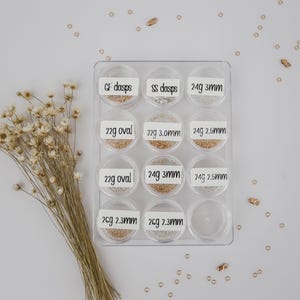 May include: A clear plastic container with 10 compartments, each containing different types of gold and silver jump rings. The compartments are labeled with the size and gauge of the jump rings.  GF dasps, SS dasps, 24g 3mm, 22g oval, 22g 3.0mm, 24g 2.5mm, 22g oval, 24g 3mm, 24g 2.5mm, 26g 2.3mm, 26g 2.3mm.