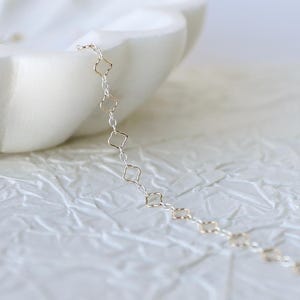 May include: A delicate silver chain necklace featuring a repeating pattern of small, open, four-petaled flower-shaped links. The metallic links are interconnected to form a continuous, slender strand that rests against a white, textured surface.