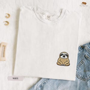 Könnte beinhalten: Ein weißes kurzärmeliges T-Shirt mit einer kleinen Stickerei eines braunen Faultiers, das mit geschlossenen Augen und offenem Mund sitzt. Das Shirt hat einen Rundhalsausschnitt und ein Etikett am inneren Kragen aus Baumwolle.