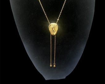 May include: A gold-colored necklace with a pendant featuring an oval design with intricate detailing. The necklace has a delicate chain and two dangling chains with small ball accents. The jewelry is displayed on a black mannequin.