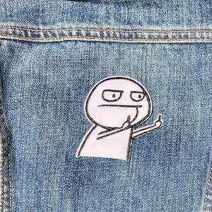 Op de afbeelding: Een witte geborduurde patch op een blauwe denim jas. De patch toont een eenvoudig cartoonfiguur met een fronsend gezicht dat de middelvinger opsteekt.