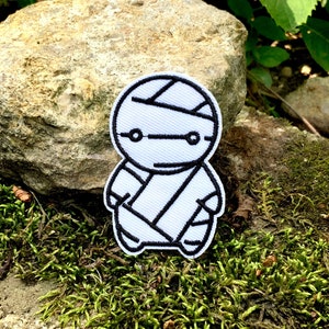 Puede incluir: Parche blanco bordado de un personaje de dibujos animados con vendas, que se asemeja a un robot, con contornos negros.