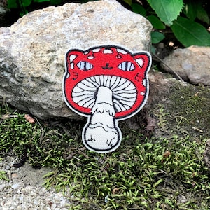 Może przedstawiać: Naszywka haftowana na czerwono i biało w kształcie grzyba z kocim pyskiem. Grzyb ma białe plamki i białą nóżkę.