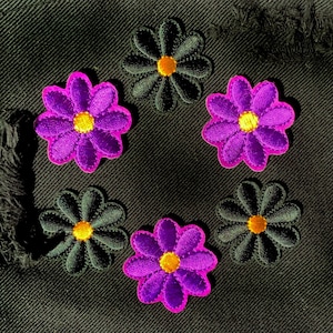 以下が含まれることがあります： 黒いデニムの背景に円形に配置された6つの刺繍された花のワッペン。花は黄色い中心を持つ紫と黄色い中心を持つ黒です。