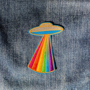May include: A rainbow-colored UFO patch with a gray saucer and a beam of light. The patch is sewn onto a blue denim jacket.