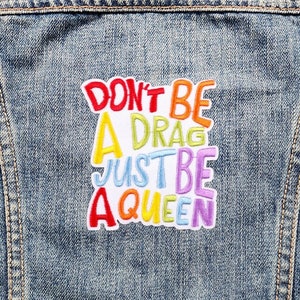 May include: A rainbow coloured embroidered patch sewn onto a blue denim jacket. The patch says "Don't be a drag, just be a queen."