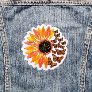 Könnte beinhalten: Ein weißer gestickter Aufnäher mit einer orangefarbenen und gelben Sonnenblume, um die herum Schmetterlinge fliegen. Der Aufnäher befindet sich auf einer blauen Jeansjacke.