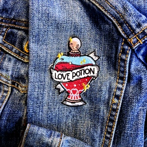 May include: A denim jacket with an embroidered patch of a heart-shaped bottle with the text "LOVE POTION" on a banner. The patch is red, white and blue.