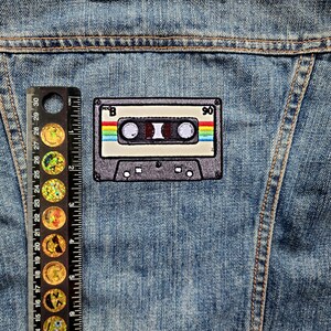 May include: A vintage-style cassette tape patch with a rainbow stripe design. The patch is sewn onto a blue denim jacket.