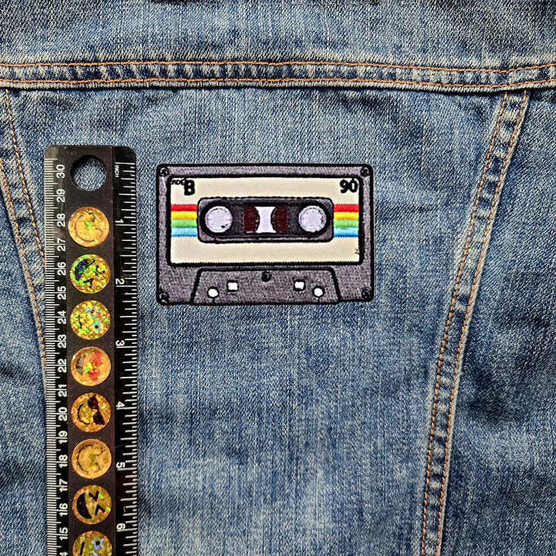May include: A vintage-style cassette tape patch with a rainbow stripe design. The patch is sewn onto a blue denim jacket.