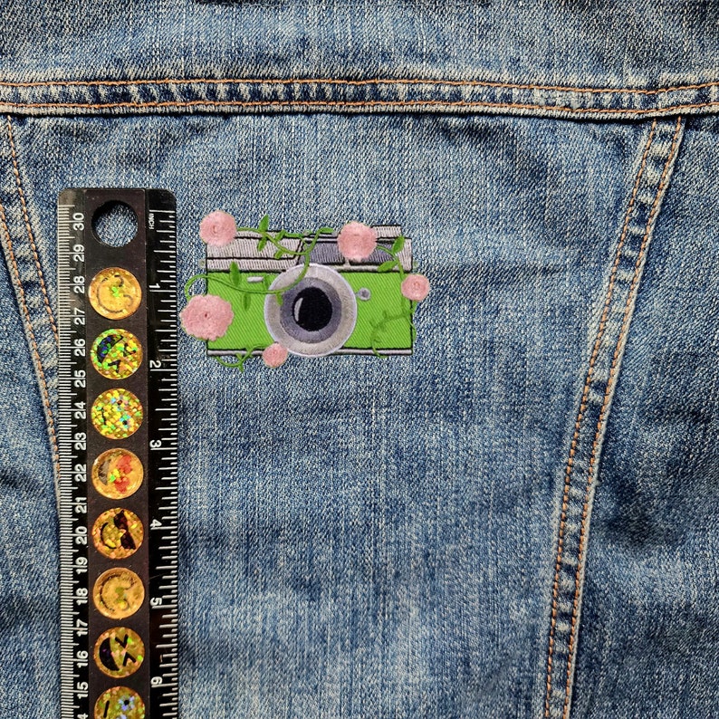 May include: A green camera with a black lens and pink flowers embroidered on a denim jacket.