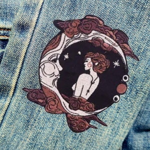 Peut inclure: Un patch brodé représentant une femme debout devant un croissant de lune avec des étoiles. La lune est entourée de nuages, le tout dans des tons de brun et de blanc.