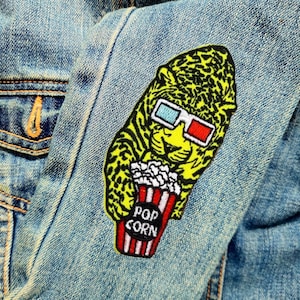 May include: A yellow and black embroidered patch of a cartoon leopard wearing 3D glasses and holding a red and white striped bucket of popcorn that says "POP CORN".