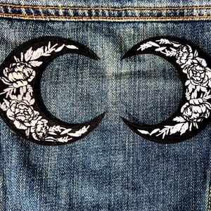 Può includere: Due toppe a forma di mezzaluna nera con ricami floreali bianchi cucite su una giacca di jeans blu.