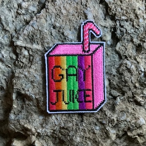 Può includere: Una toppa ricamata a forma di scatola di succo, con un design a strisce arcobaleno e il testo "gay juice".