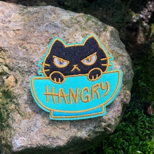 Puede incluir: Un gato negro con ojos enojados asoma por el borde de un tazón turquesa con la palabra "HANGRY" bordada en hilo dorado.