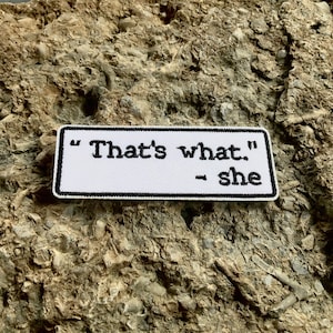 May include: White fabric patch with black embroidered text that reads "That's what." - she. The patch is on a rough, gray, textured surface.